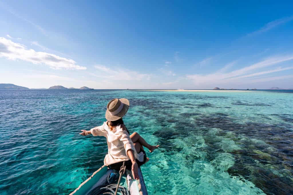 Traveler sitting on the bow of a wooden vessel, gliding over crystal-clear coral reefs during a serene private Komodo boat trip.